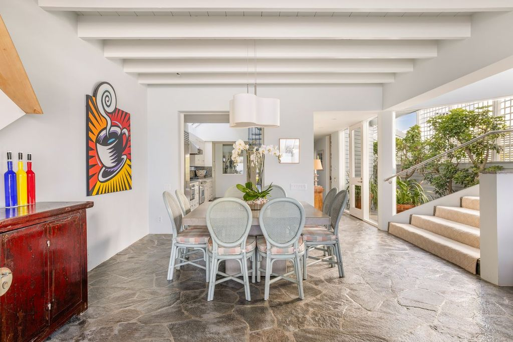 A rectangular table with woven chairs fills the center of the room, while open doorways on either side connect it to the kitchen and a planted courtyard.