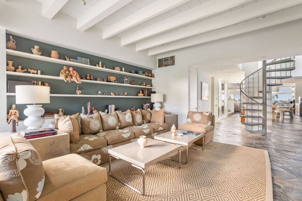 A floral-patterned sectional and matching armchair gather around a pair of square tables, while long built-in shelves stretch across the back wall to display books, pottery, and sculptural pieces against a muted green backdrop.