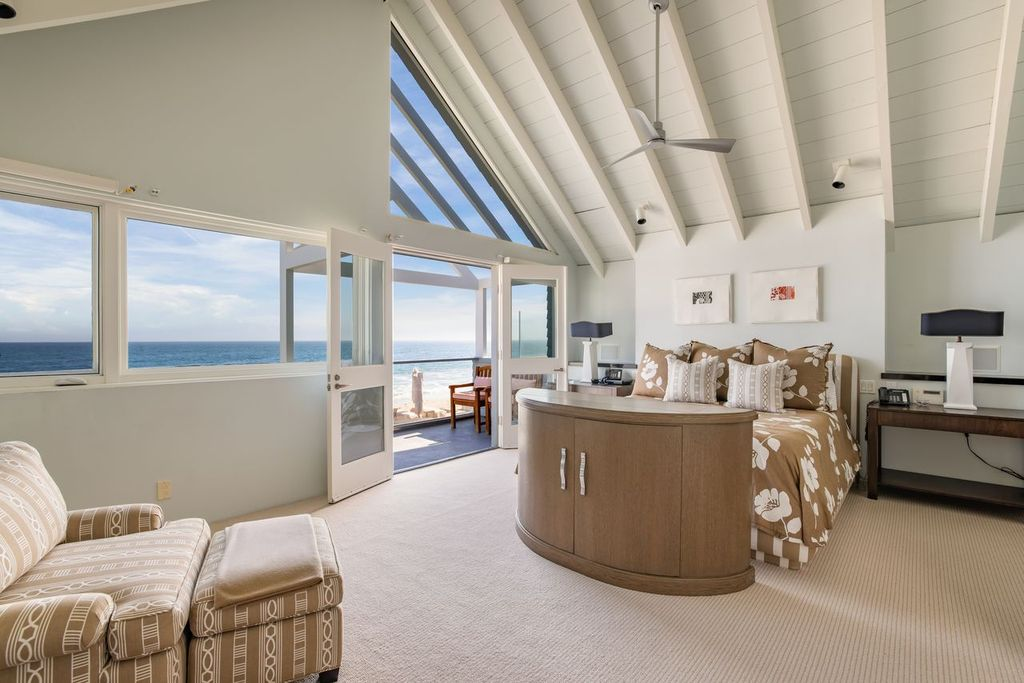 The bed is positioned behind a curved cabinet at the foot, while wide windows and double doors open the room directly toward the water.