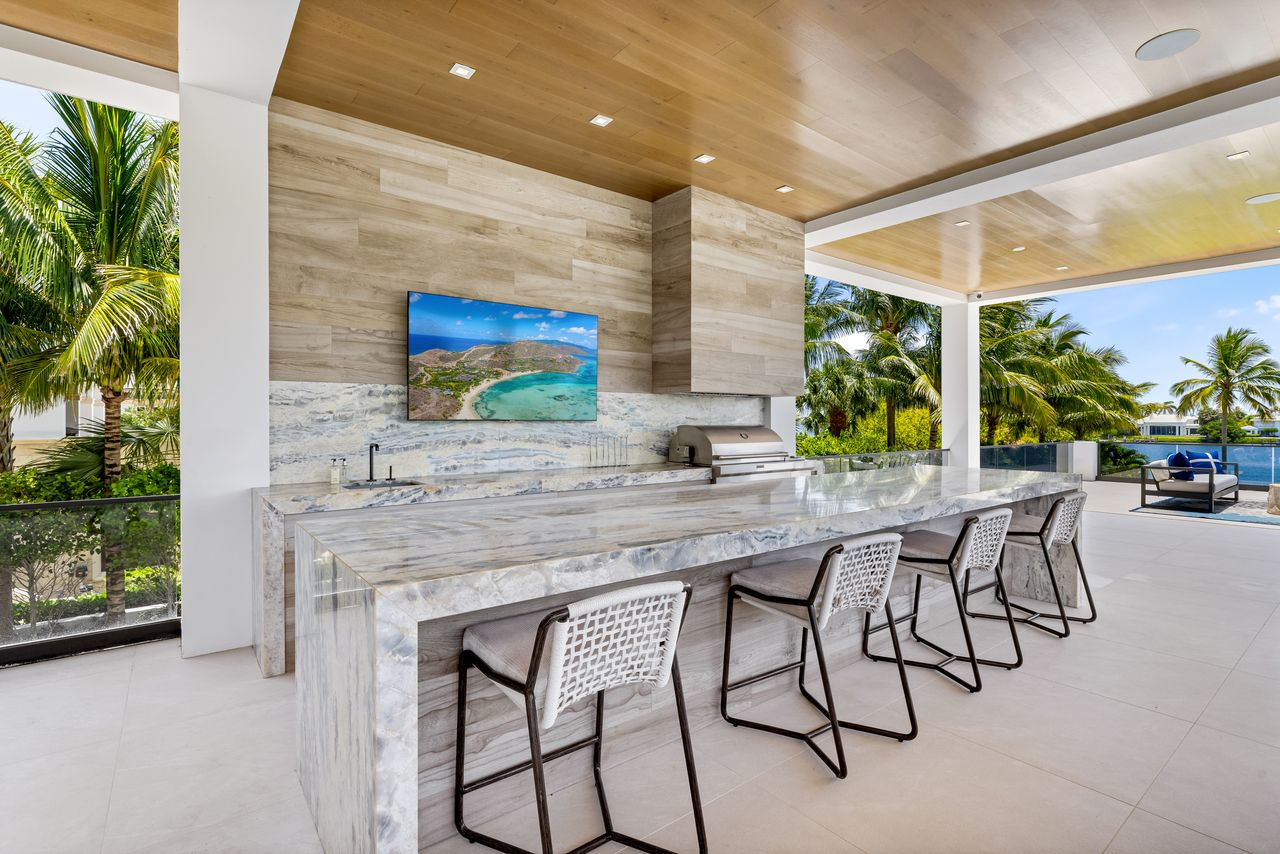 One side of the loggia features a fully equipped summer kitchen finished with dramatic waterfall stone countertops that wrap around an oversized island with bar seating.