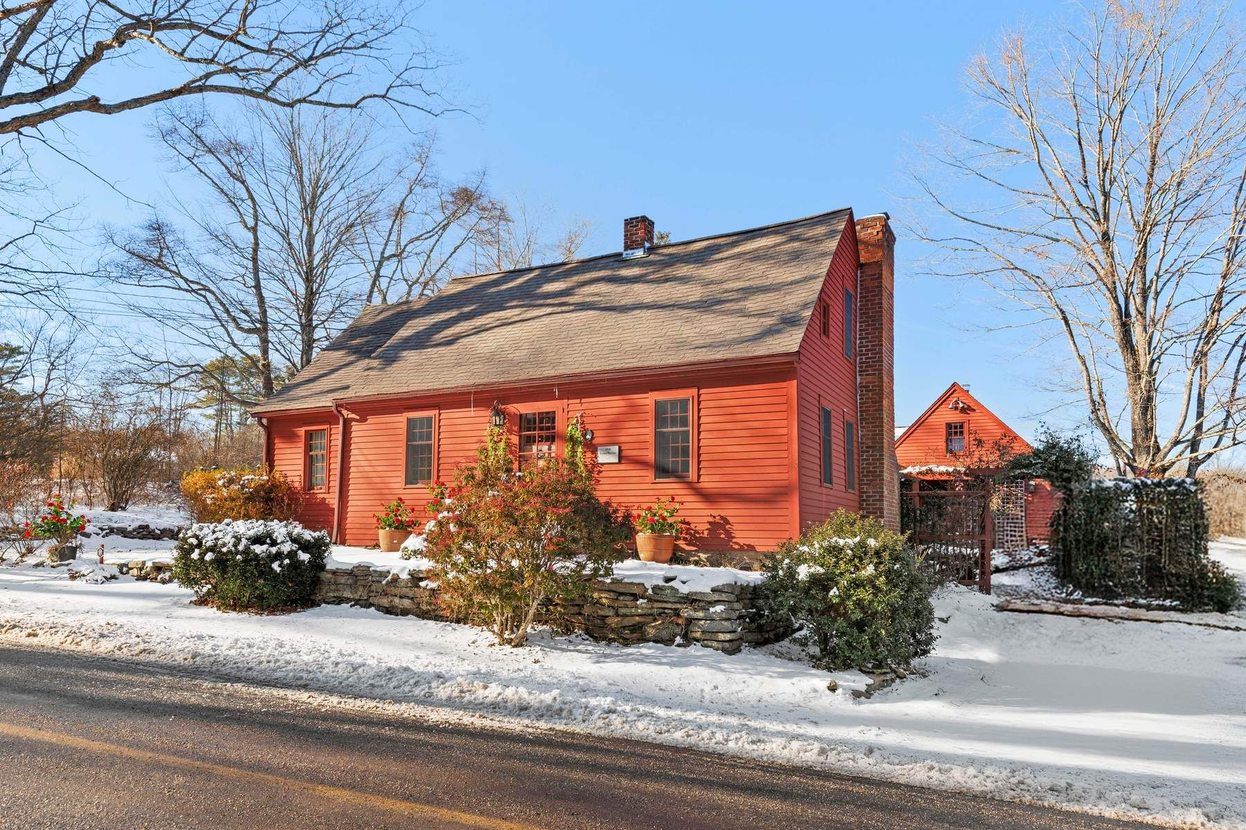 A timeless New England exterior with red clapboard siding, brick chimneys, and a steeply pitched roof framed by bare trees and stone-lined gardens.