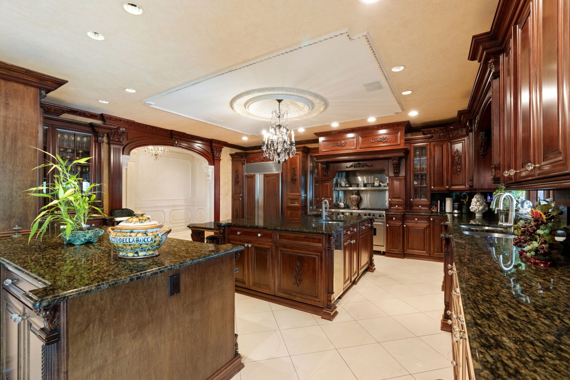 Eat-in kitchen is wrapped in carved-wood cabinetry and dark stone countertops, with a large central island providing additional prep space and storage beneath another crystal chandelier.
