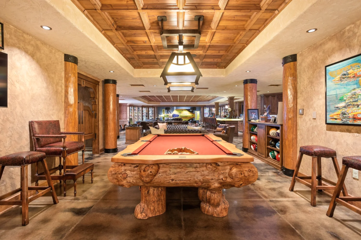Around the corner from the bar, the entertainment space continues into a billiards area, where a live-edge-style pool table sits beneath a large light fixture and another coffered wood ceiling.