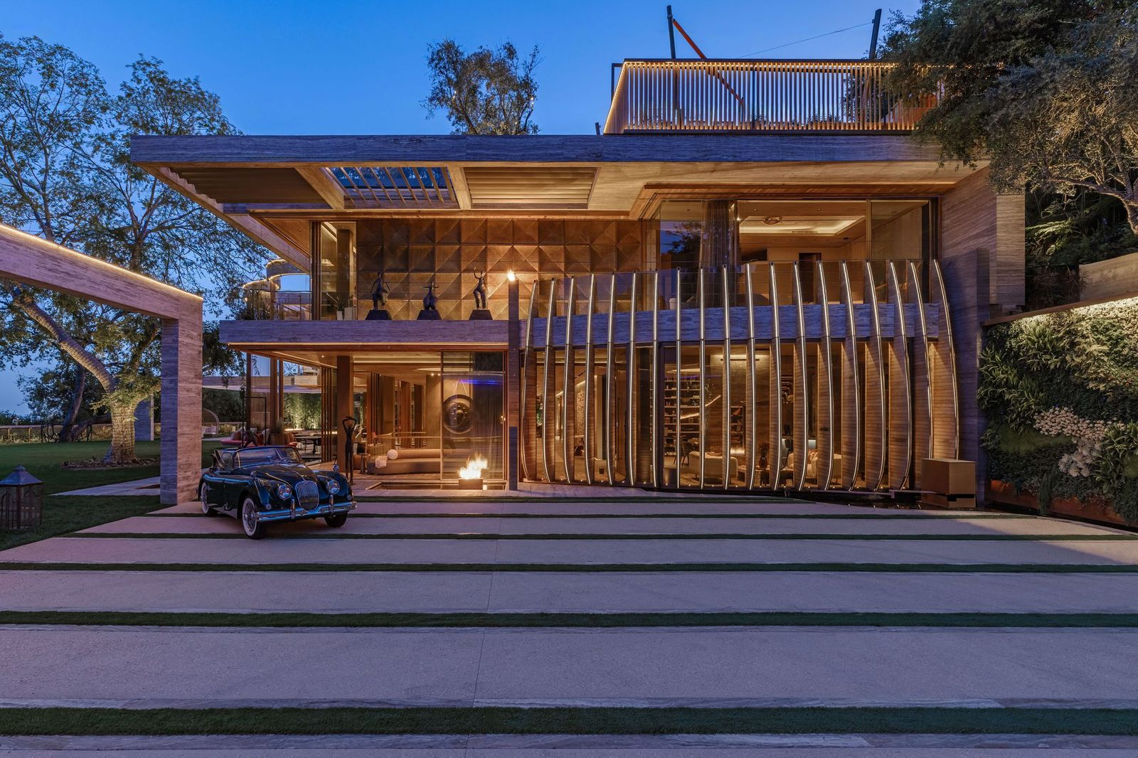 Tall stone walls, timber screens, and tall greenery frame the front of the house behind a broad concrete driveway.