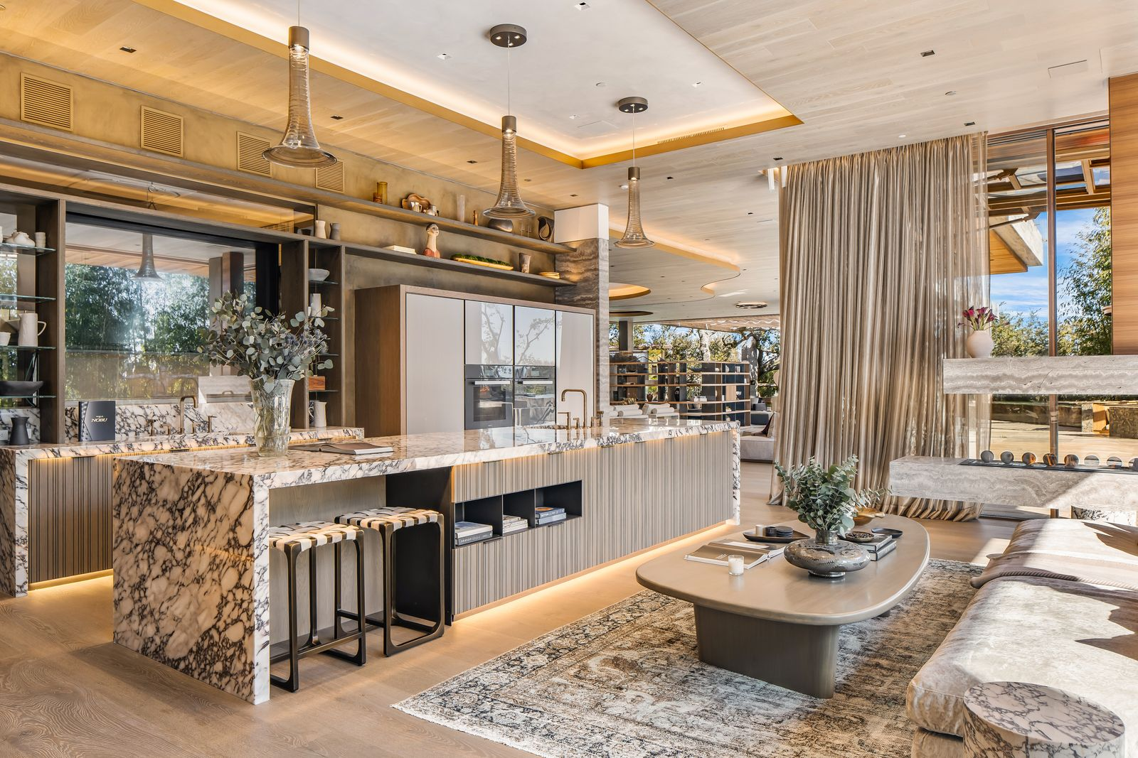 Pendant lights drop over the work surface, while pale wood ceilings, bronze-toned cabinetry, and layered stone keep the room visually connected to the rest of the main floor.