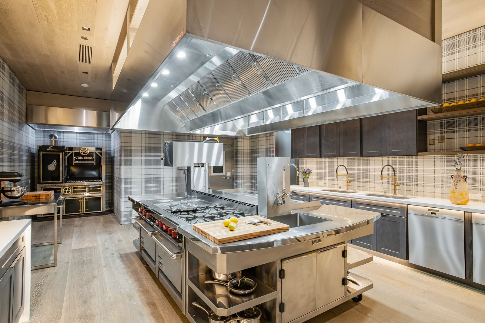A large stainless-steel cooking island fills the center beneath an oversized exhaust hood, with professional ranges, ovens, refrigeration, and prep sinks arranged around the perimeter.