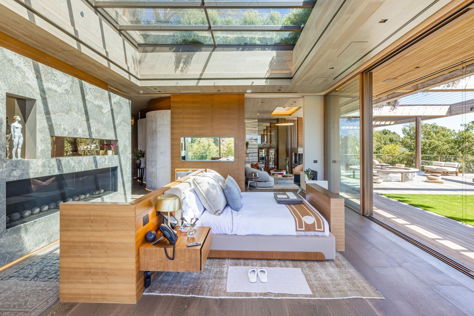 Warm wood panels frame the bed, while a long stone fireplace runs along the back wall and floor-to-ceiling openings pull the surrounding trees and sky into view.