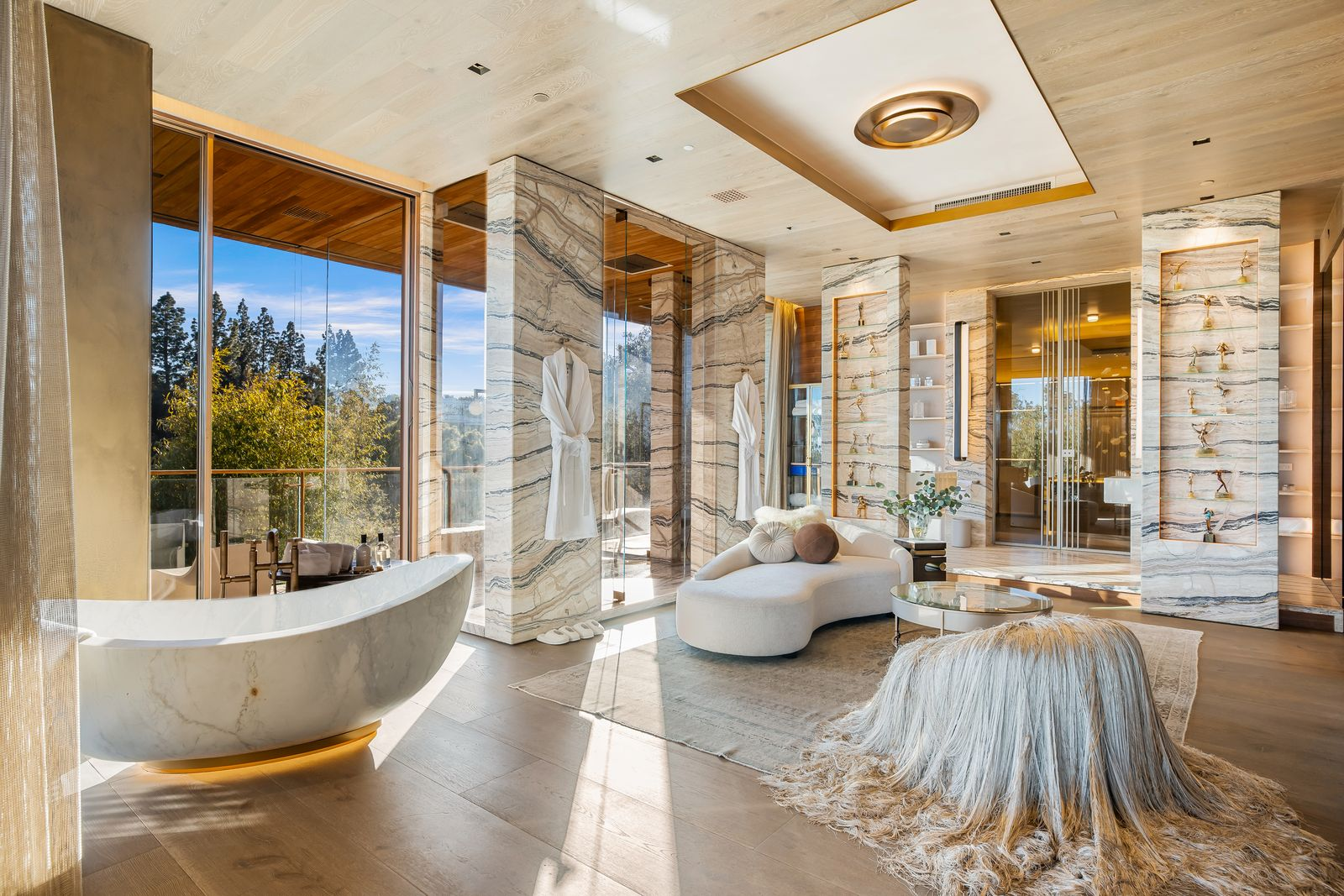 The suite's bathroom is lined in veined stone, pale wood, glass, and bronze detailing, with a freestanding stone tub that sits beside the windows and a glass-enclosed shower.
