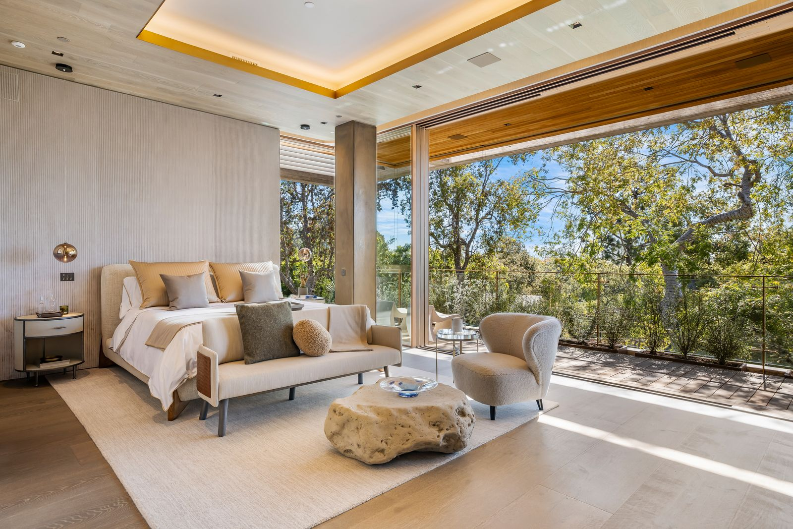 A low upholstered bed sits against a softly textured wall, with a small sofa and sculptural stone coffee table creating a sitting area at the foot of the bed.