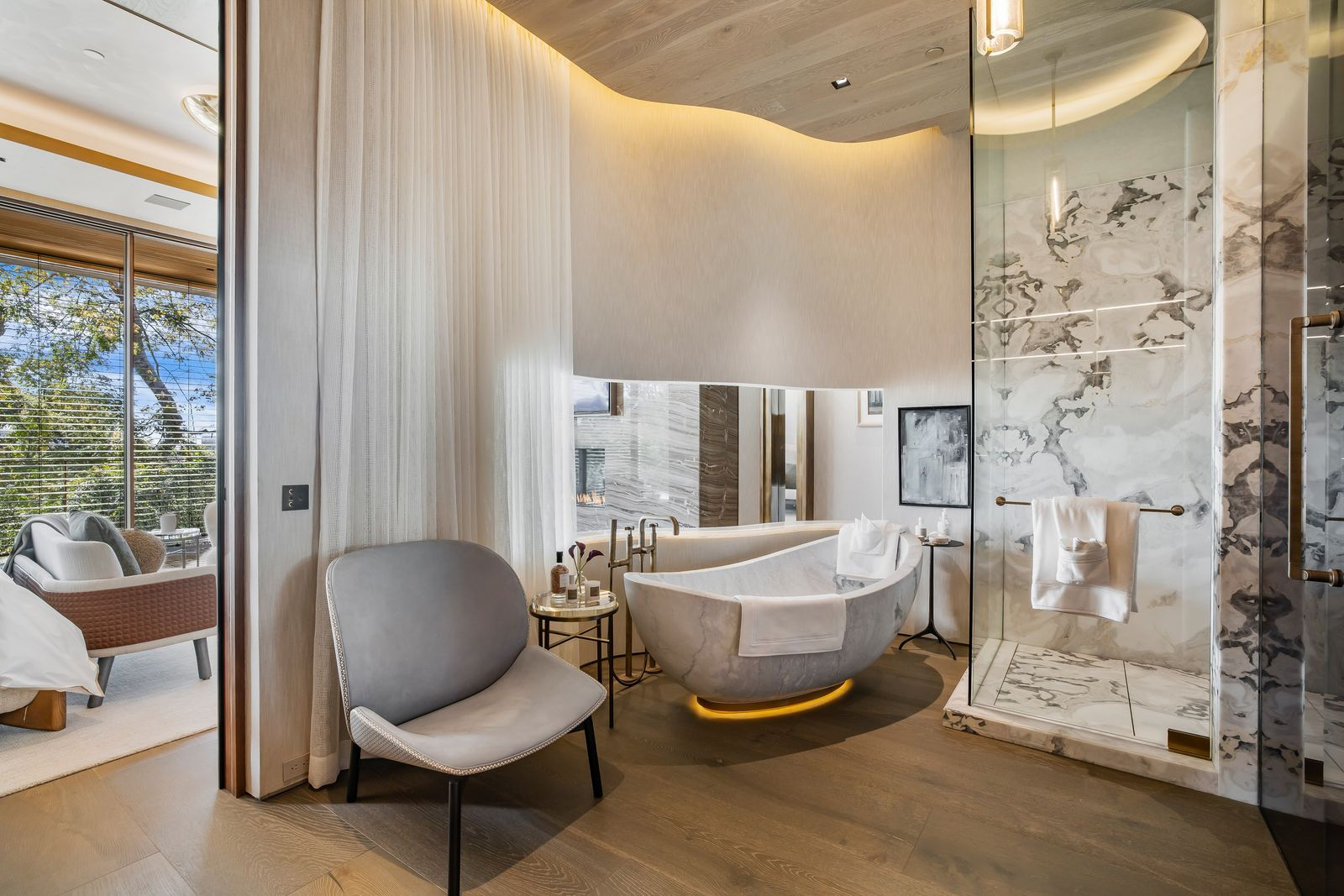 The en-suite bathroom continues the lighter palette with a freestanding stone tub, a glass shower wrapped in veined marble, and curved architectural lines that soften the transition between the bedroom and bath.