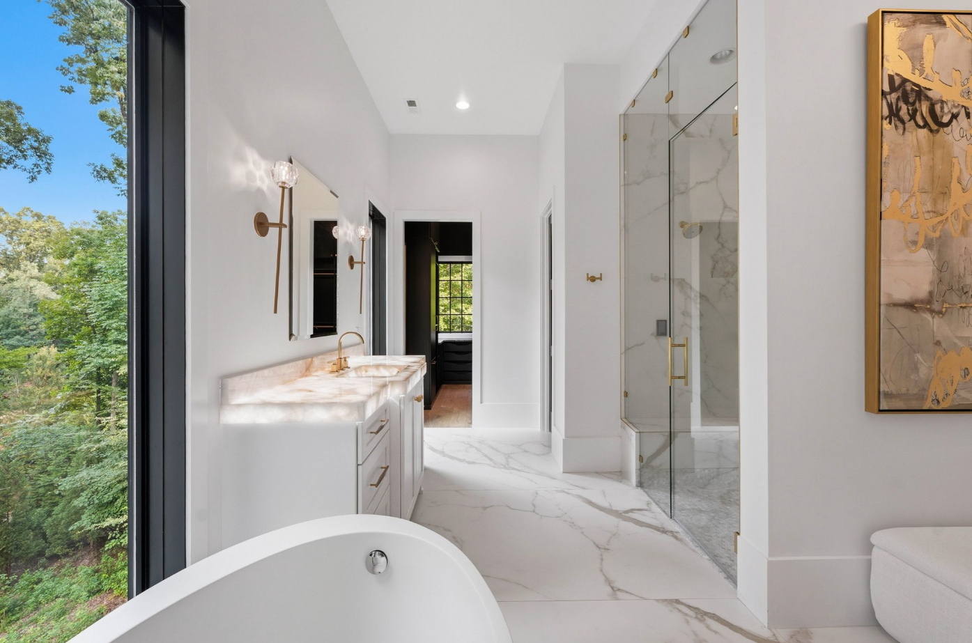 A freestanding soaking tub is positioned beside a large window, while dual vanities extend along either side of the shower with backlit stone counters, brass fixtures, and individual mirrors.