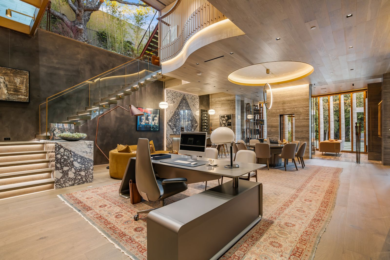 At the bottom of a staircase, the home office opens as a large, gallery-like workspace with a desk in the foreground and a round meeting table positioned farther into the room.