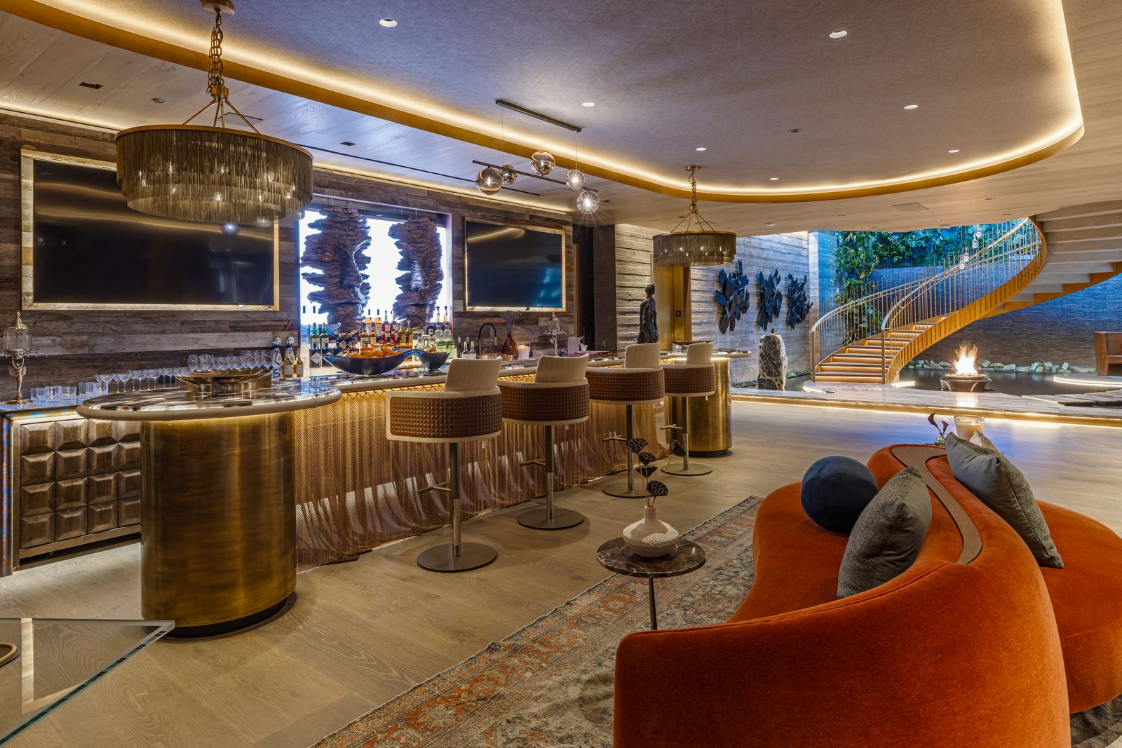 At the bottom of the bronze spiral staircase is the social lounge centered around a bronze bar. The curved counter is lined with stools and backed by illuminated shelves of bottles and glassware.
