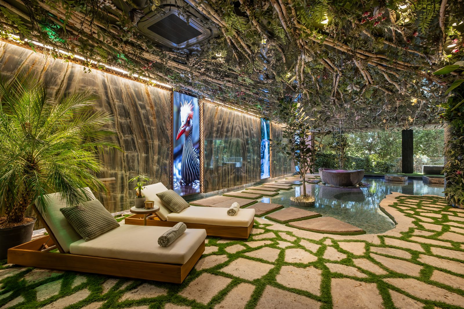 Stone paths weave around the water, while lounge chairs sit beneath a ceiling densely covered with branches and greenery.