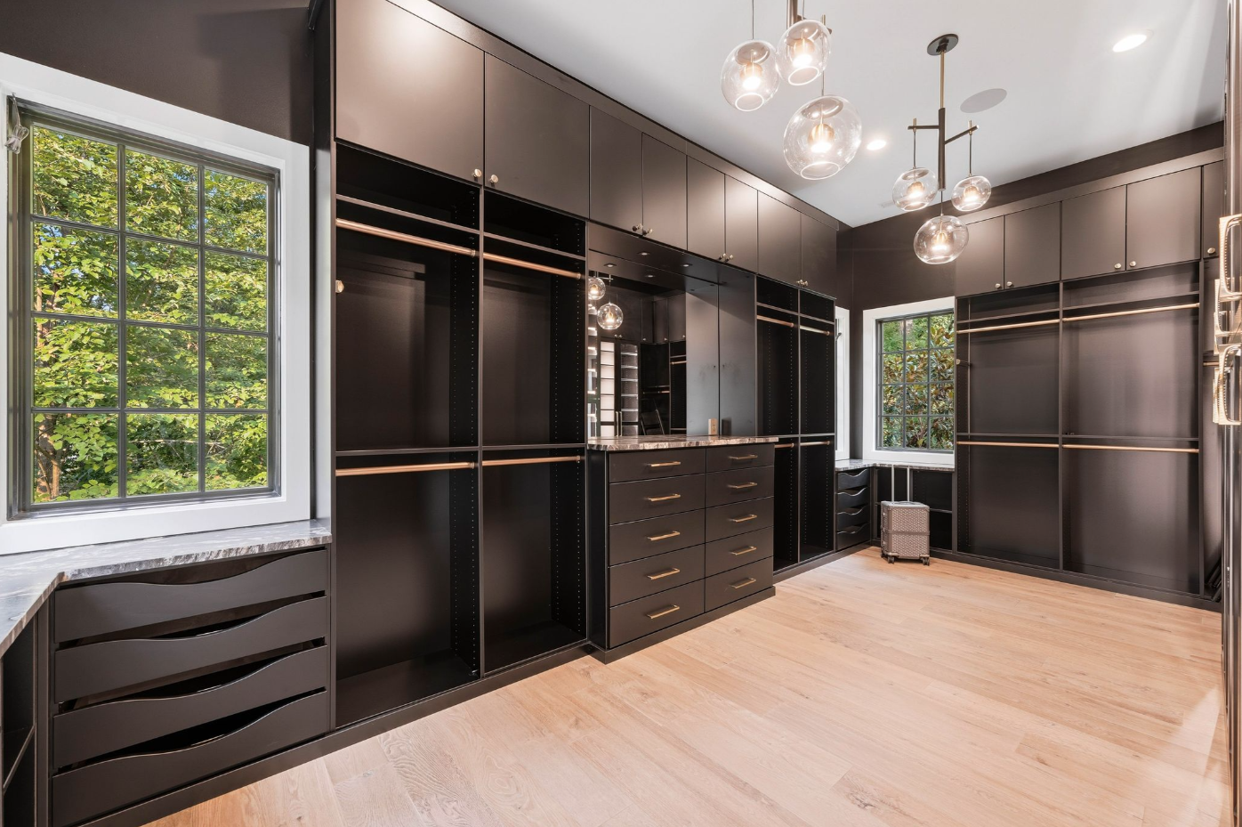 Tall windows bring natural light into the dark millwork, while glass-front wardrobes, illuminated shelving, and sculptural pendant fixtures give the spaces the layout of a private boutique.
