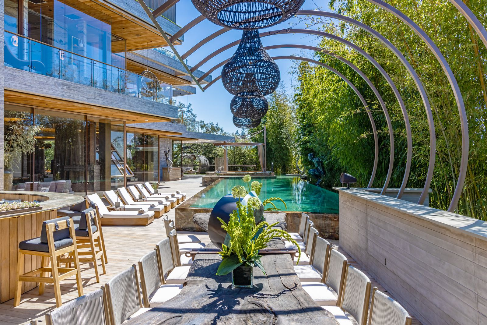 A wood table runs through the center beneath a series of curved metal ribs and oversized woven pendant fixtures, with a bar positioned alongside it.