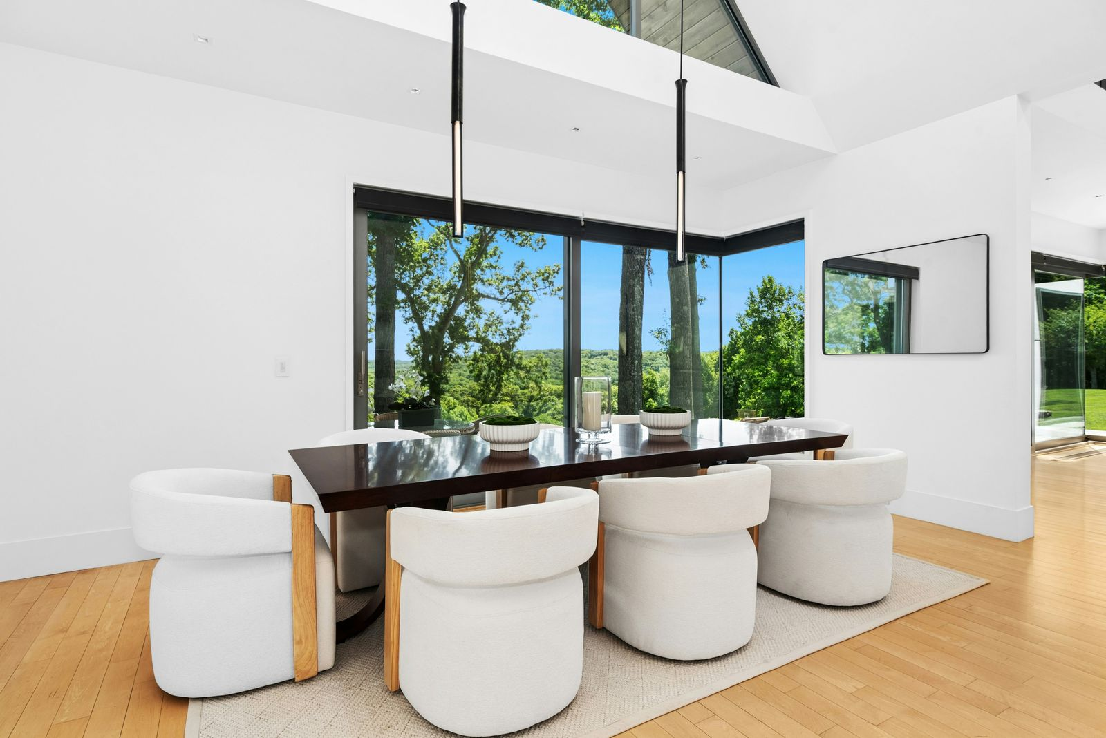 A dining space with a long dark wood table surrounded by rounded upholstered chairs set beneath two slim pendant lights that drop from the vaulted ceiling.