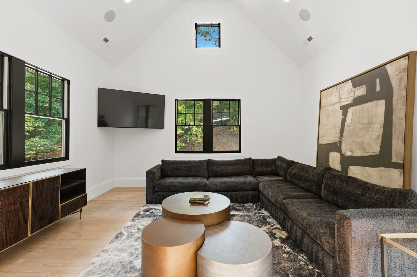 A charcoal sectional follows the corner of the room beneath black-framed windows, while overlapping brass-toned tables and oversized artwork give the space a casual media-lounge layout.