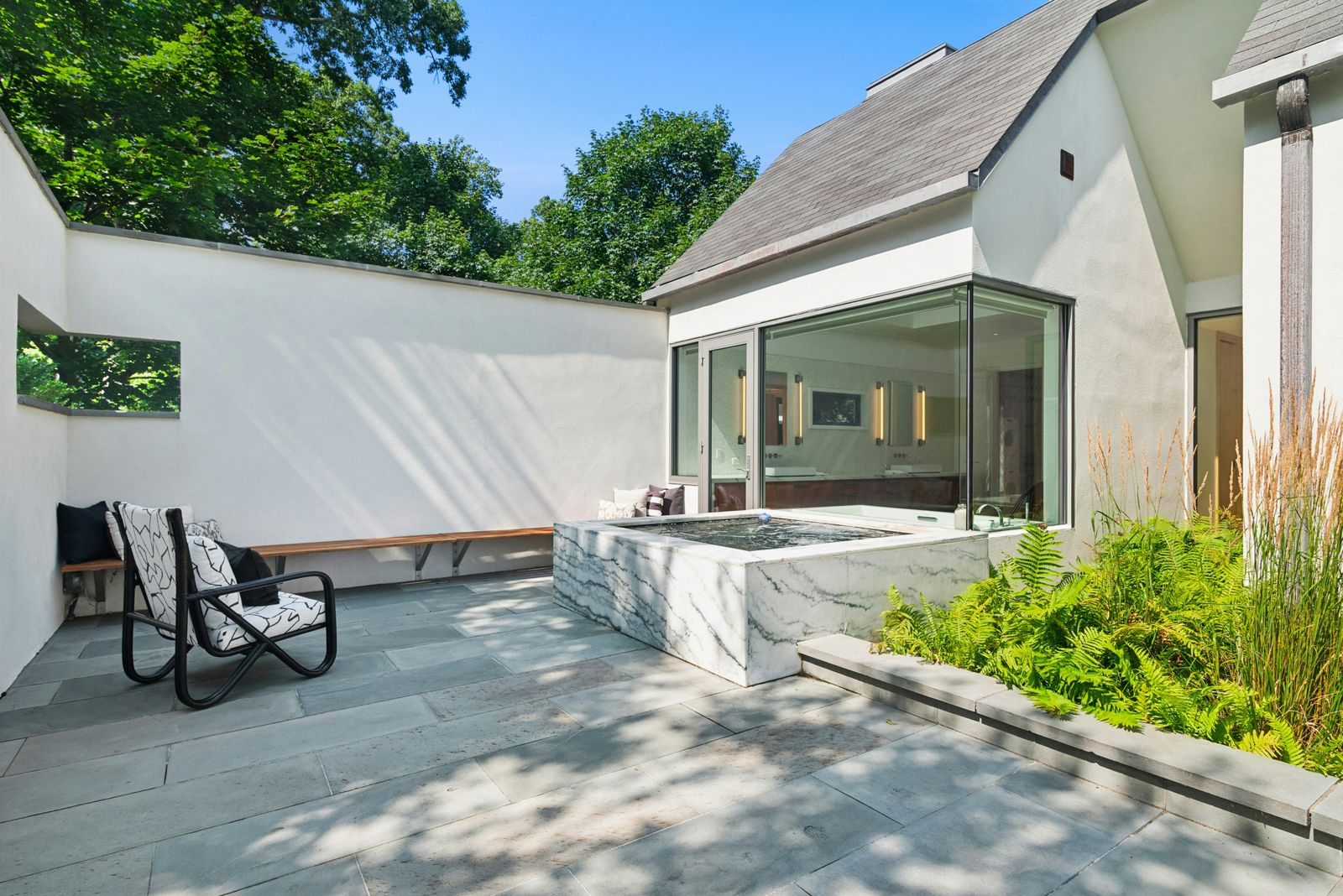 A square marble hot tub along the bathrooms walls, with built-in bench seating along the perimeter and planted ferns softening one side of the courtyard.