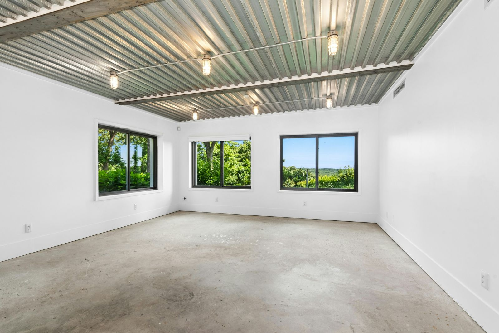 The industrial-style artist’s studio with a raw concrete floor and an exposed corrugated-metal ceiling crossed by steel beams and simple utility lighting.