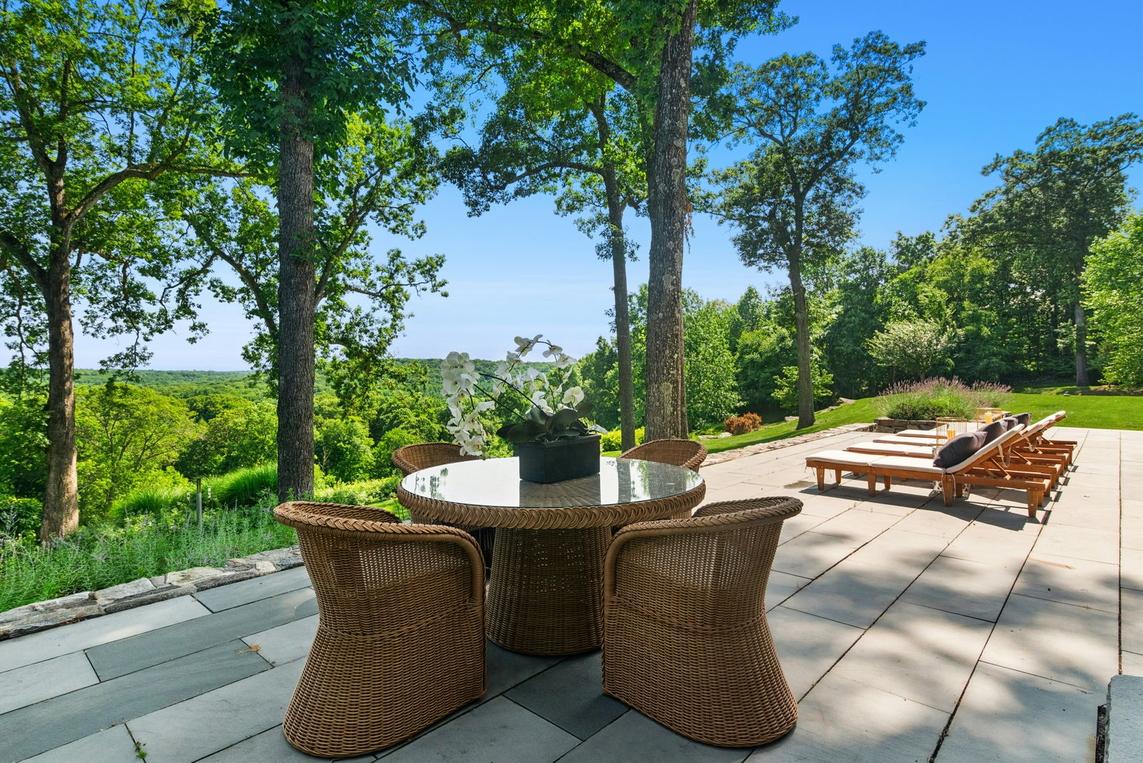A round dining table occupies the edge of the terrace, where the paved surface gives way to landscaped slopes and views across the surrounding tree canopy.
