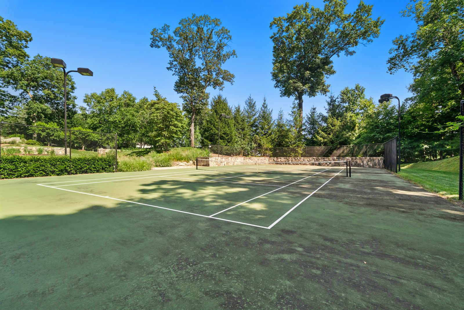 A full-size lighted tennis court occupies its own clearing within the trees surrounded by fencing and stone retaining walls.