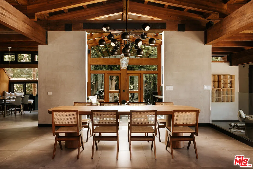 At the center, a long wood dining table is surrounded by cane-backed chairs beneath a sculptural black chandelier.