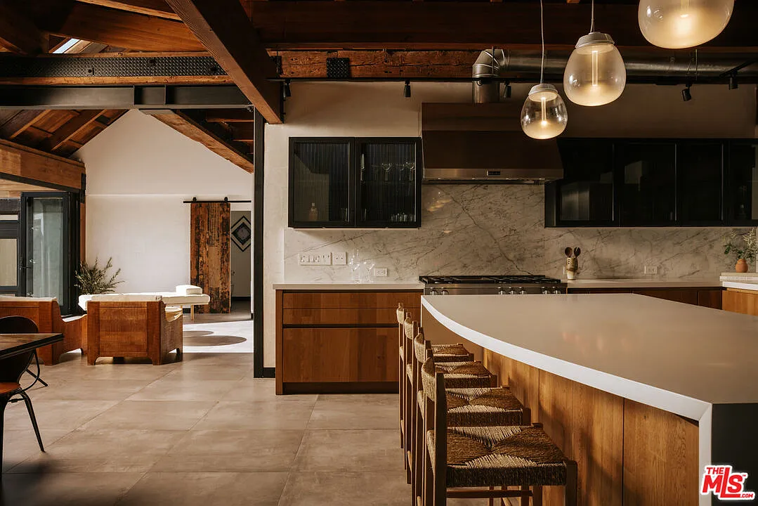 Along the perimeter, more wood cabinetry is paired with white stone counters and a marble backsplash, while dark upper cabinets frame the range and oversized hood.