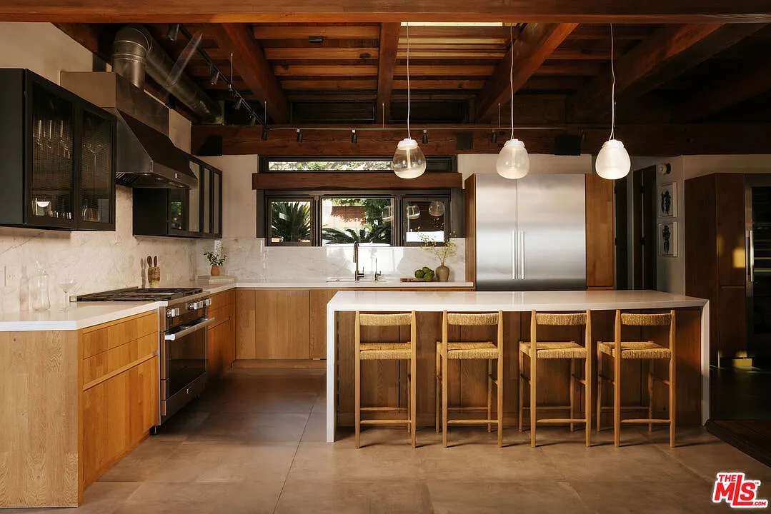 The kitchen is built around a long island with seating for four, its white countertop wrapping around warm wood cabinetry beneath a row of glass pendant lights.