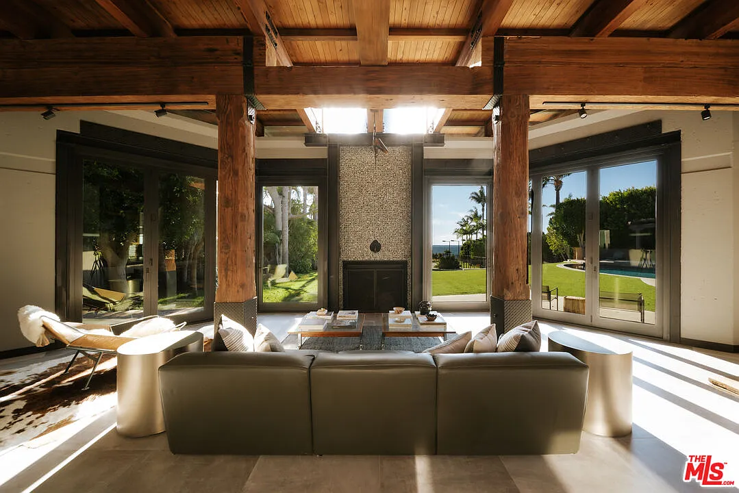 Heavy timber posts and beams frame the room, while large glass doors and windows wrap the outer walls with views toward the lawn, pool, and ocean.