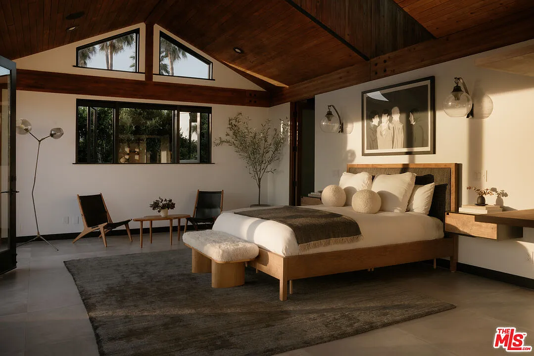 The primary suite opens beneath a vaulted wood ceiling, with the bed set against one wall and a small sitting area arranged near the opposite side.