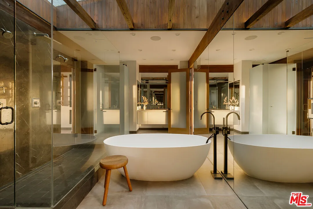A freestanding oval tub sits at the center of the room between a horizontal glass-enclosed shower and a long mirrored wall.