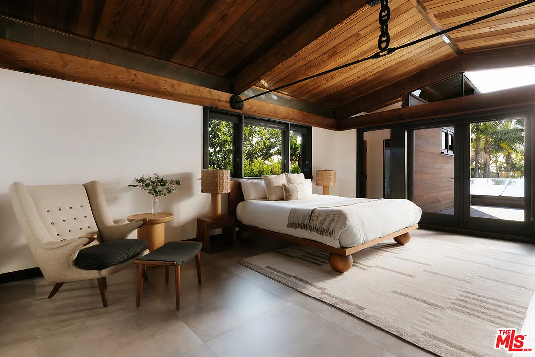 The first bedroom sits beneath a steeply pitched wood ceiling with a low wood-framed bed and a small sitting chair placed before the terrace doors.