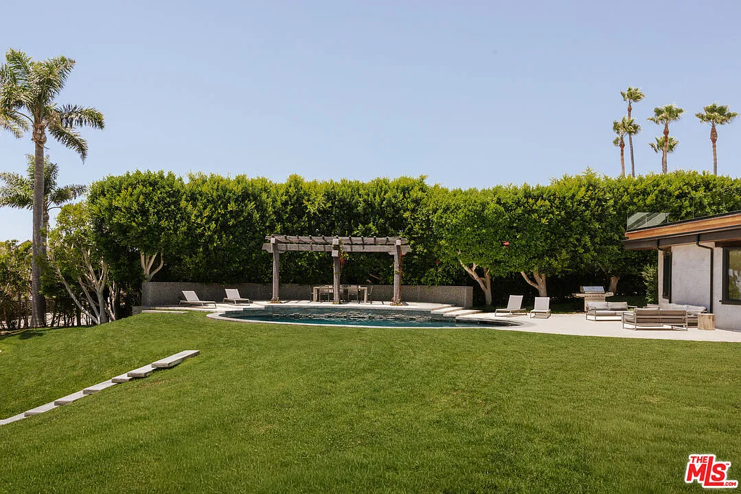 A curved swimming pool is surrounded by a broad stone patio with lounge seating and space for outdoor dining.