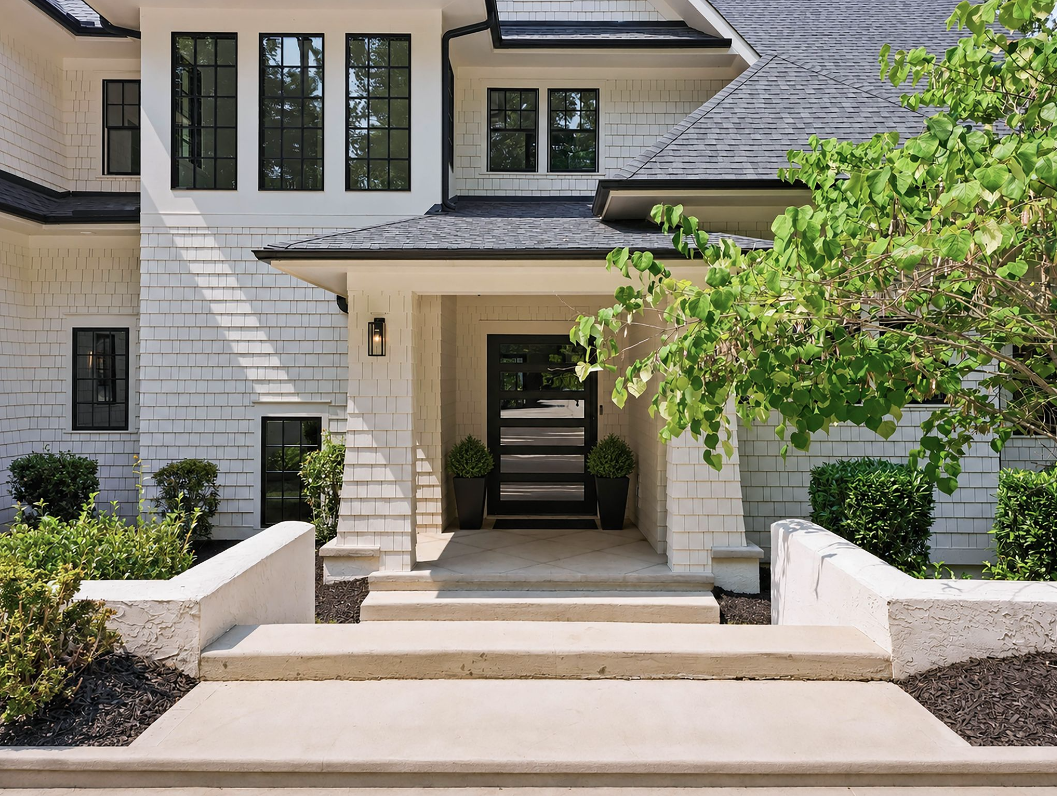 At the center of the residence, a short stairway guides you beneath a covered entry to a large pivoting front door, framed by black-trimmed windows and white shingle siding.