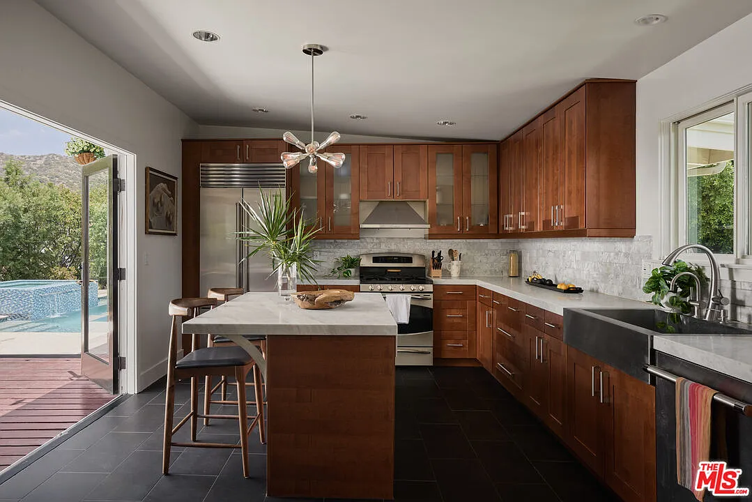 The kitchen is wrapped in rich wood cabinetry on three sides, paired with pale stone counters, a lightly textured backsplash, stainless-steel appliances, and a farmhouse-style sink.