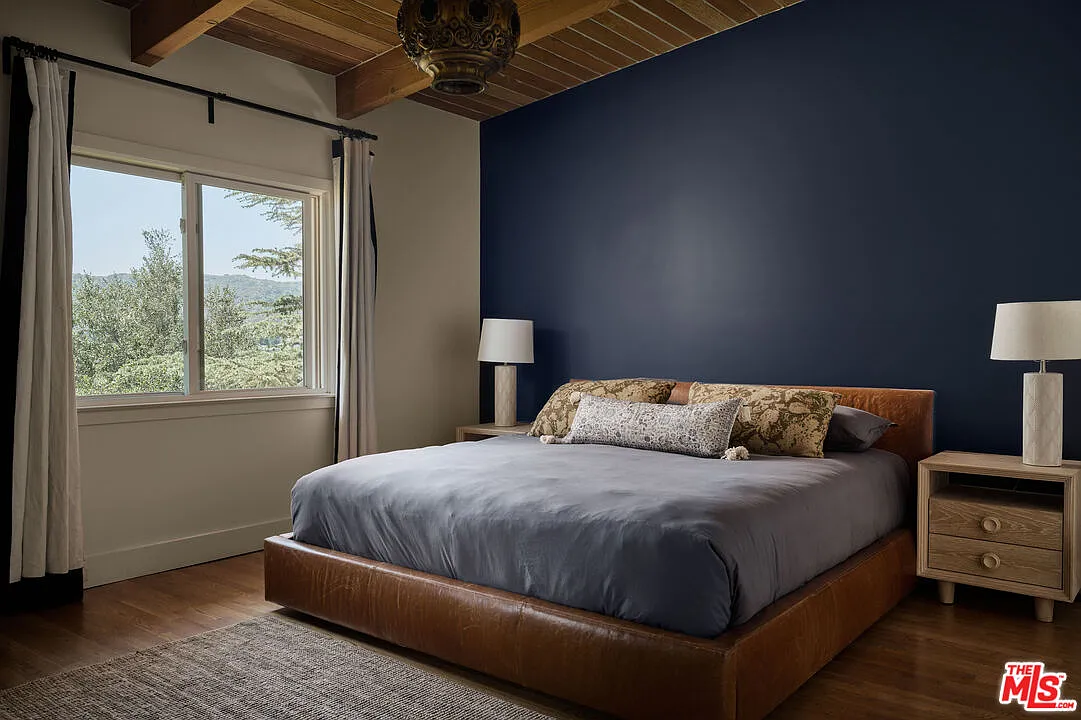 The primary suite continues the home’s mid-century palette with exposed ceiling beams, warm wood floors, and a deep blue accent wall behind the bed.