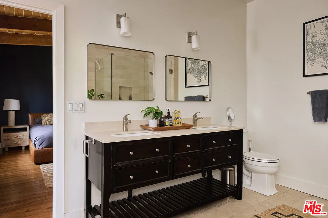 The connected bathroom has a much brighter palette, pairing pale stone tile with a long dark-wood double vanity and twin mirrors.