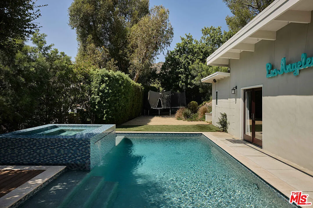 The swimming pool sits between a tall privacy wall of greenery and the music studio with a raised, mosaic-tiled hot tub positioned along one side.