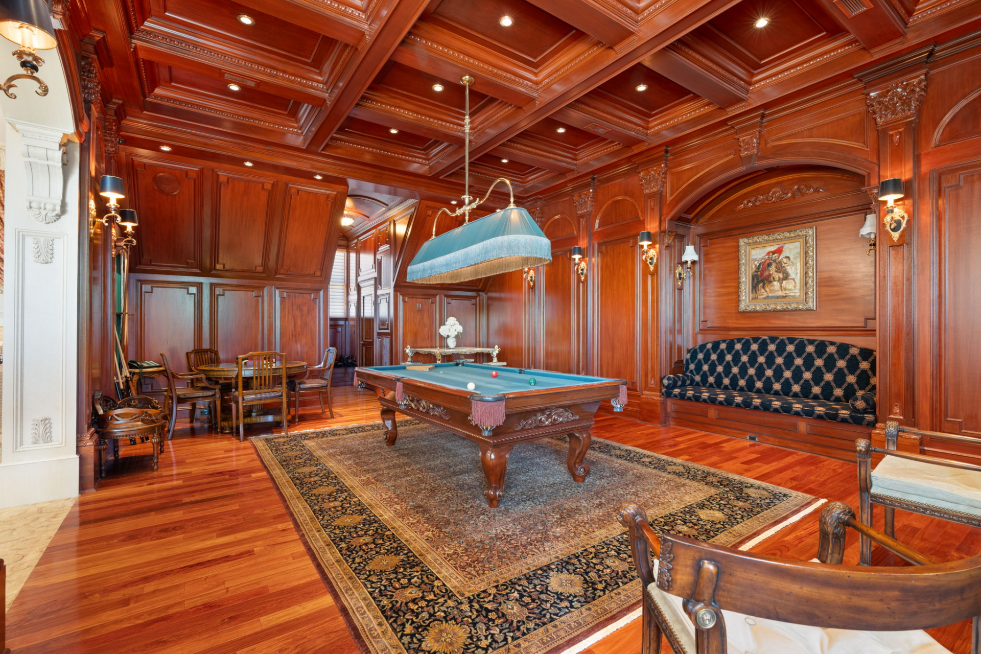A billiards room wrapped in rich wood paneling that covers the walls and coffered ceiling, creating a club-like setting centered on a carved pool table beneath an oversized light.