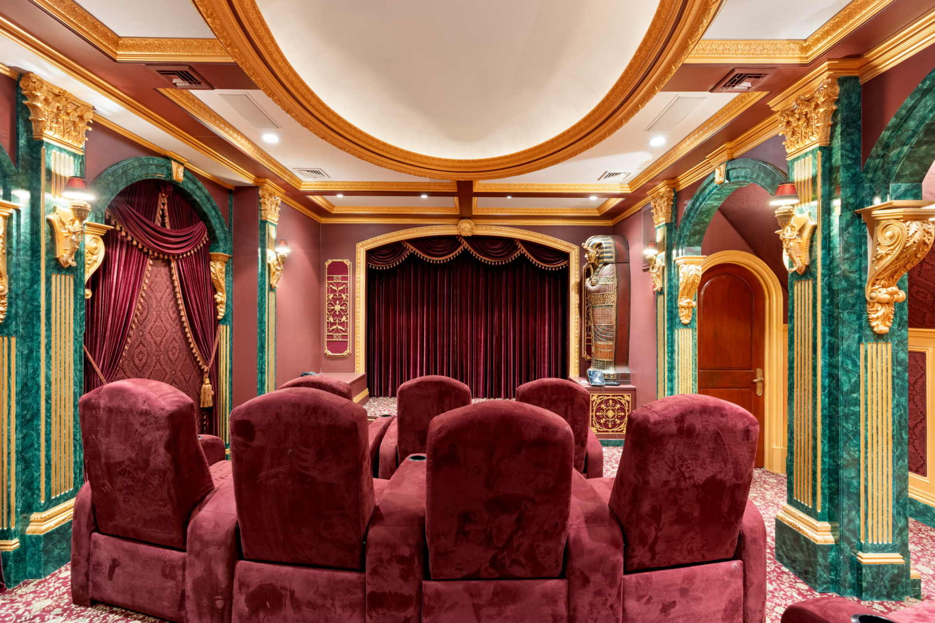 The movie theater features rows of deep burgundy reclining seats facing a curtained screen, while green marble columns, gold trim, and red walls surround the room.