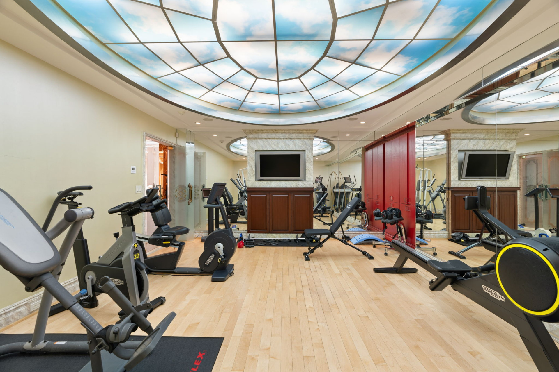 A dedicated fitness room filled with cardio machines, free weights, benches, and strength-training equipment arranged beside a full mirrored wall.