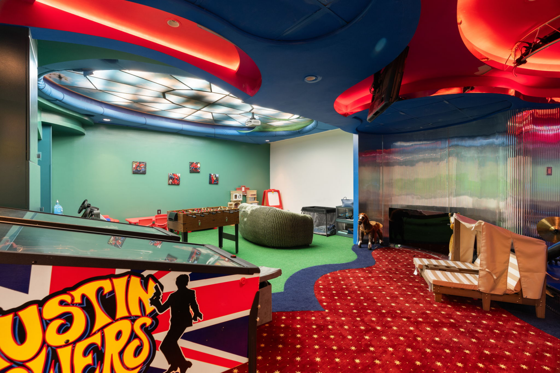 A colorful arcade and play space with pinball machines, foosball, lounge seating, TVs, and additional kids' games beneath a sculpted blue-and-red ceiling.