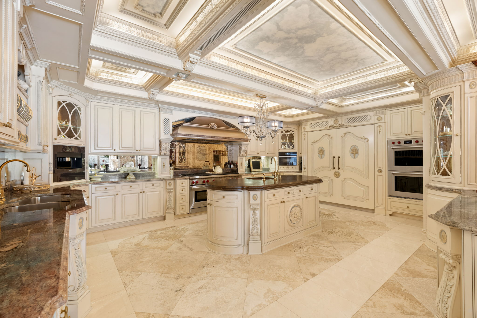 The room is arranged around a central island with extensive surrounding cabinetry, multiple sinks, professional-grade appliances, and stone work surfaces.