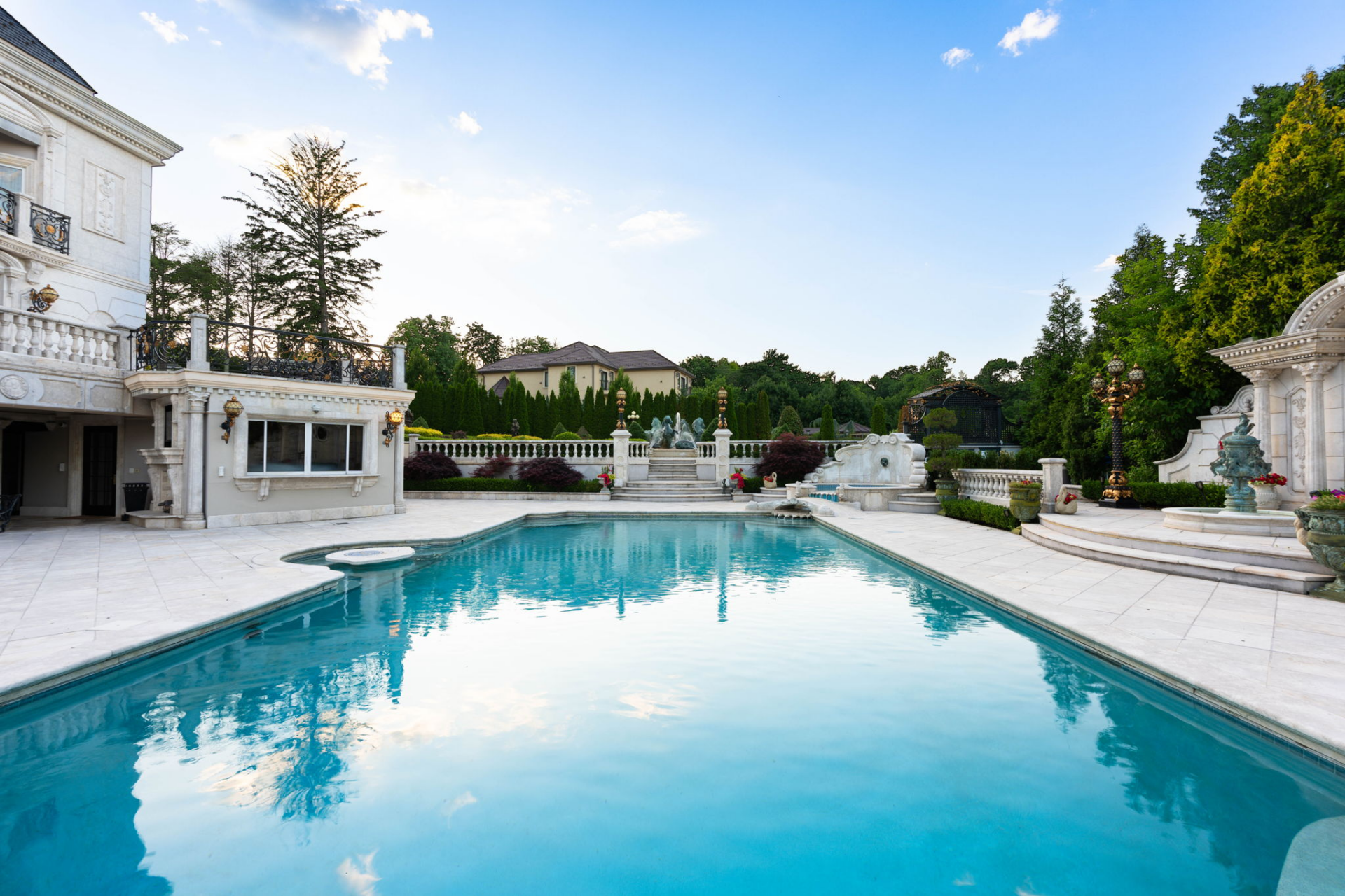 The pool itself is framed by wide expanses of pale stone paving, with a raised spa built into one corner and a separate outdoor bar positioned nearby for serving the surrounding seating areas.