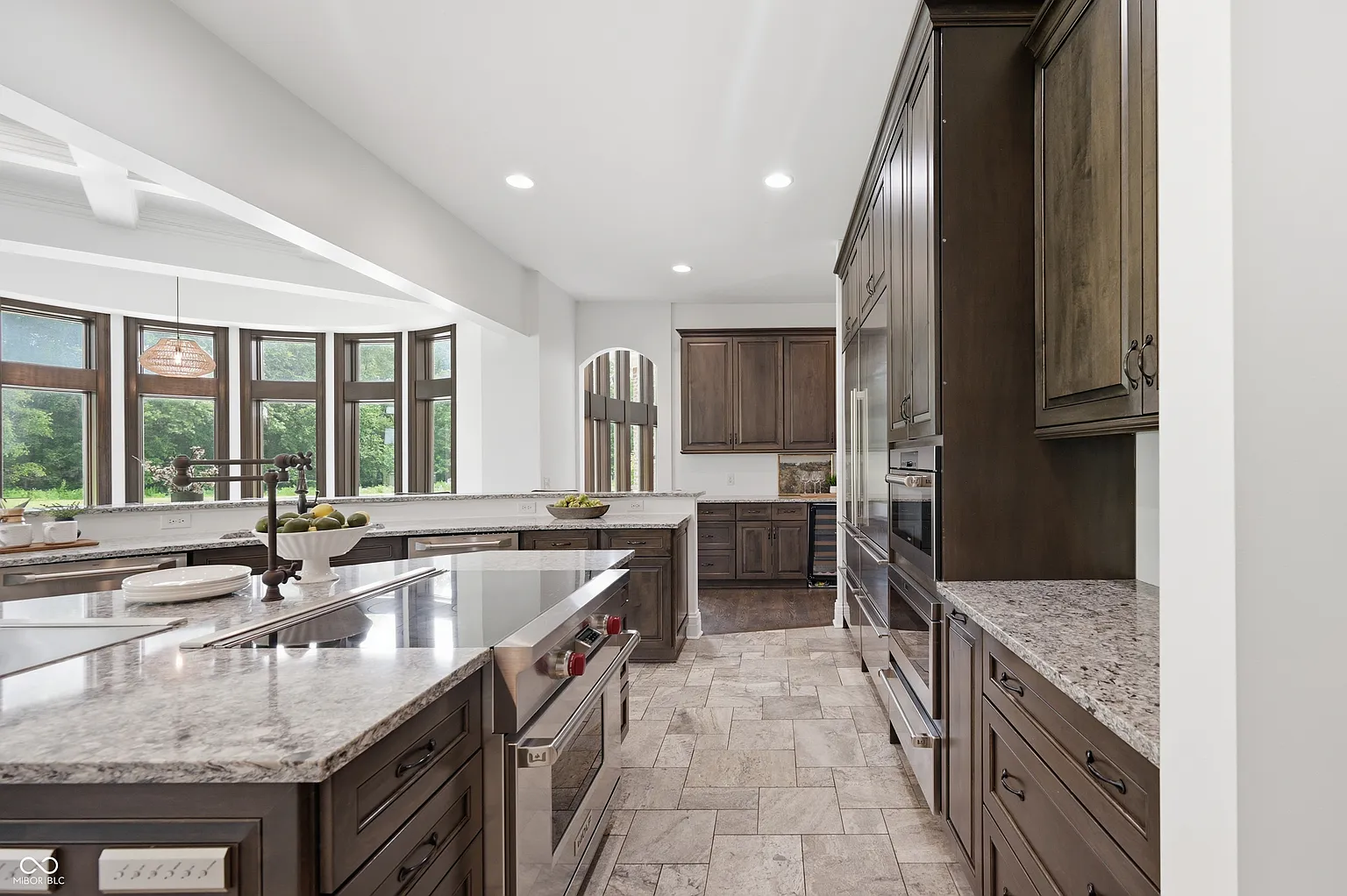 Rark wood cabinetry extends across the walls around a built-in stainless-steel refrigerator, stacked wall ovens, and a dishwasher. 