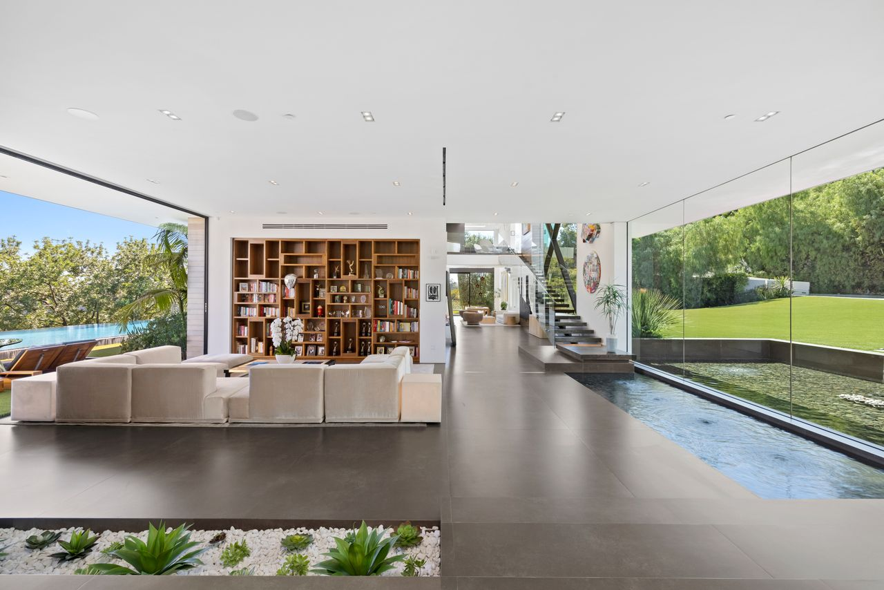 Stepping inside, the home opens to a soaring atrium where glass walls, dark stone floors, and shallow reflecting pools surround a sunken sitting area.