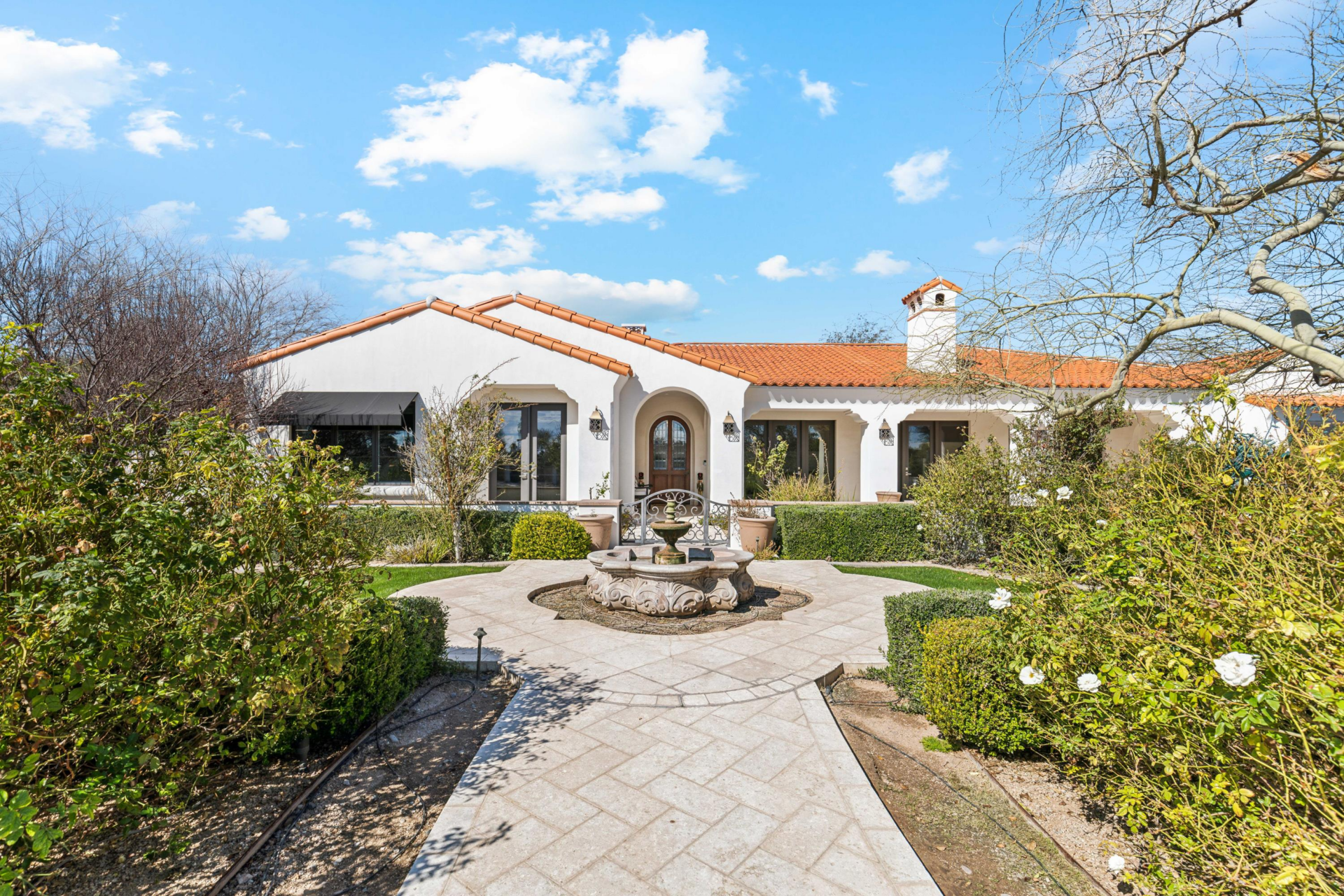 A paved walkway cuts through the landscaped front grounds before arriving at a circular fountain courtyard framed by hedges and flowering plants.