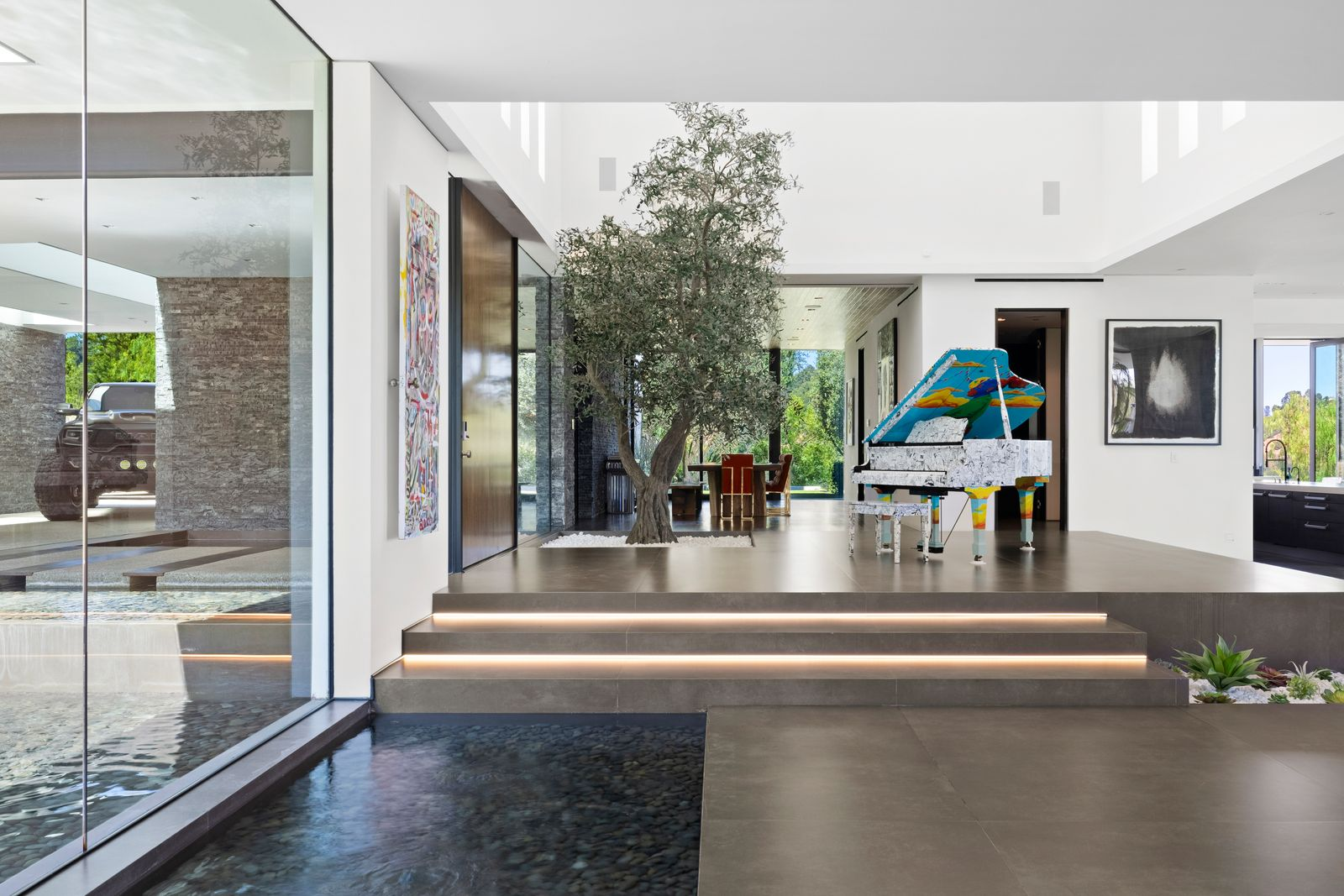 Illuminated steps rise to the next level of the open plan, where an indoor olive tree and a brightly painted grand piano stand beneath the double-height ceiling. 