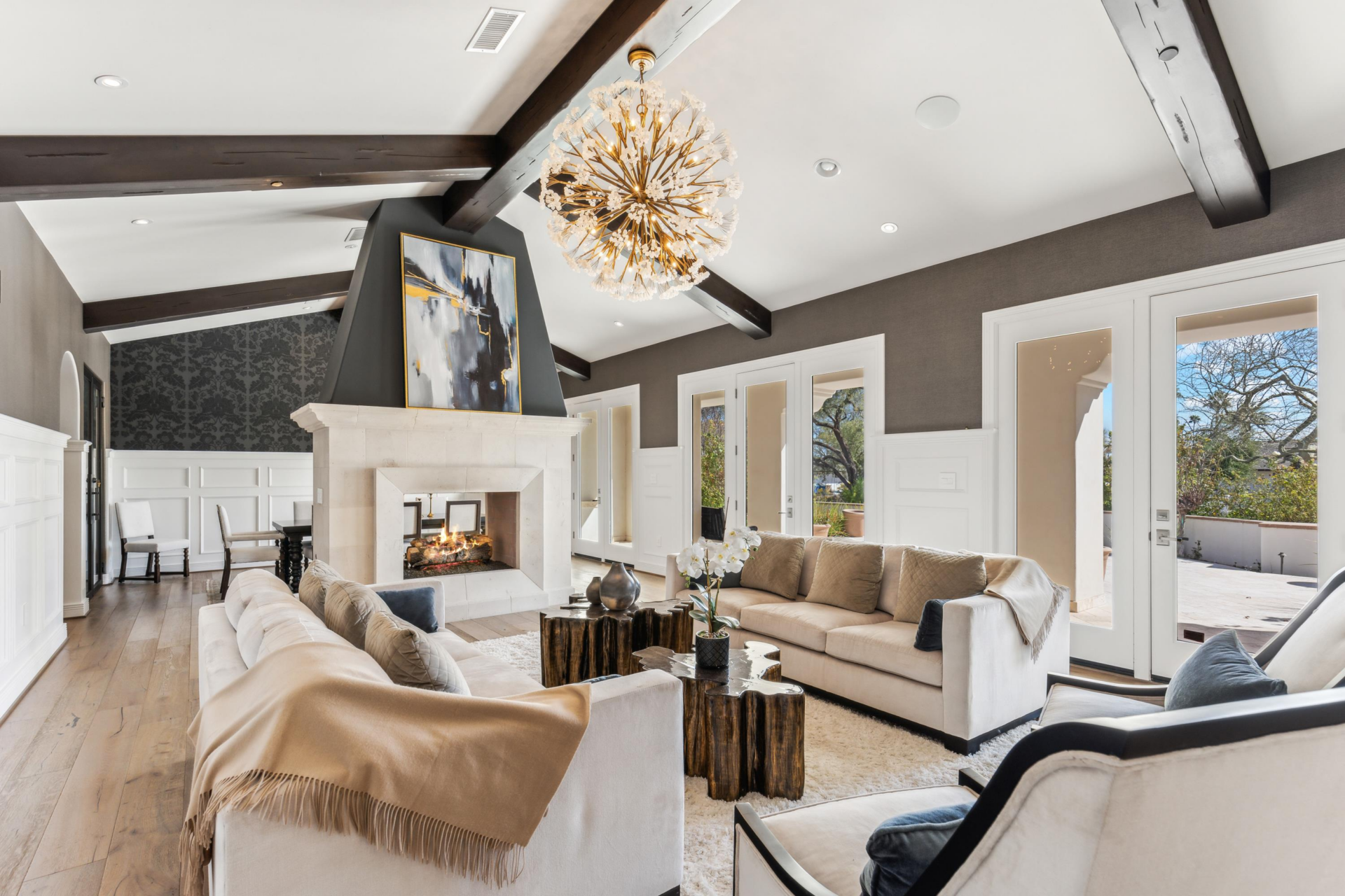 The formal living room stretches beneath a vaulted ceiling crossed by dark exposed beams, with imported hardwood floors running below a seating area arranged around the fire.